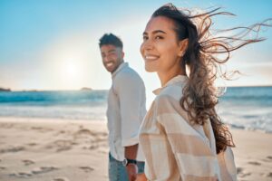 Windswept man and woman holding hands walking on the beach smiling